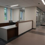 Reception desk in a modern office lobby with dark wood cabinets and a white marble-pattern panel, two black mesh chairs, and large windows to the left showing greenery outside.