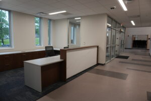 Reception desk in a modern office lobby with dark wood cabinets and a white marble-pattern panel, two black mesh chairs, and large windows to the left showing greenery outside.