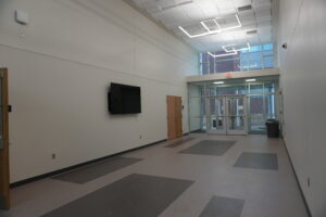 Long white-walled hallway with a patterned gray floor, a wall-mounted TV, and glass exit doors at the far end under an EXIT sign. Outside brick buildings are visible through windows.