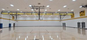 Wide view of an empty gymnasium with a basketball hoop at each end, polished wooden court, and white walls trimmed in yellow and blue.