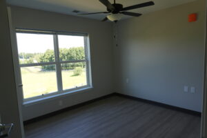 Empty, unfurnished room with gray walls, wood-look flooring, a ceiling fan, and a large window showing a green landscape outside.