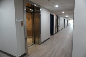 Open elevator doors in a modern hallway; a wall sign reads 'ELEVATOR' beside the doors, with a long corridor to the right.