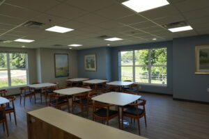 Bright classroom or lounge with white tables and wooden chairs, large windows letting in greenery outside.