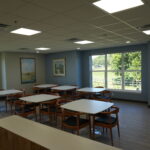 Large bright classroom or teaching room with white rectangle tables and wooden chairs, blue-gray walls, and large windows showing trees outside.