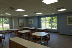 Large bright classroom or teaching room with white rectangle tables and wooden chairs, blue-gray walls, and large windows showing trees outside.