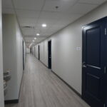 Long clinical corridor with gray walls, bright ceiling lights, and a dark blue door on the right side of the hall.