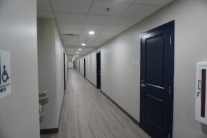 Long clinical corridor with gray walls, bright ceiling lights, and a dark blue door on the right side of the hall.