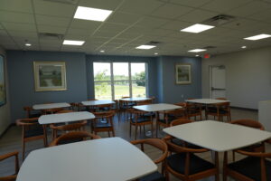 Bright classroom or dining area with white rectangular tables and wooden chairs, blue walls, and large windows showing a green landscape outside.