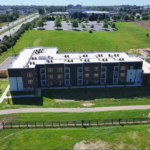 Aerial view of a modern three-story apartment building with brick and dark panels, set in green fields; a highway to the left and a parking lot behind.