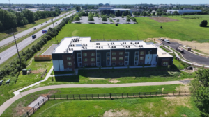 Aerial view of a modern three-story apartment building with brick and dark panels, set in green fields; a highway to the left and a parking lot behind.