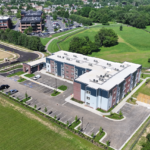 Aerial view of a modern U-shaped apartment building with blue-gray panels, surrounding parking and green fields nearby.