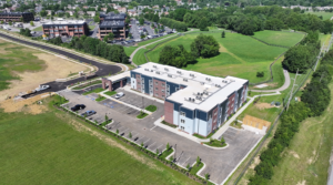 Aerial view of a modern U-shaped apartment building with blue-gray panels, surrounding parking and green fields nearby.