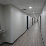 Long, well-lit office hallway with gray walls, a water fountain on the left, and doors along both sides of the corridor.
