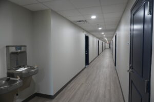 Long, well-lit office hallway with gray walls, a water fountain on the left, and doors along both sides of the corridor.