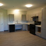 Bright modern kitchen with gray upper and lower cabinets, white countertops, a stainless steel stove and range hood opposite a sink row, and a doorway on the left.