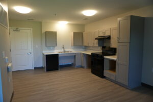 Bright modern kitchen with gray upper and lower cabinets, white countertops, a stainless steel stove and range hood opposite a sink row, and a doorway on the left.