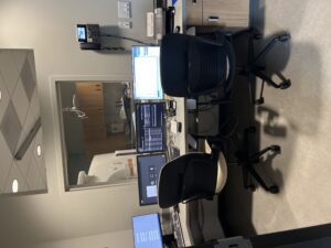 Office workspace with several computer monitors and two chairs; doorway opens to a room with cabinets and medical imaging equipment beyond.