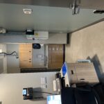 Small medical office or clinic room with wooden cabinets, sink, and countertop along back wall; ceiling-mounted equipment hangs overhead.