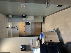 Small medical office or clinic room with wooden cabinets, sink, and countertop along back wall; ceiling-mounted equipment hangs overhead.