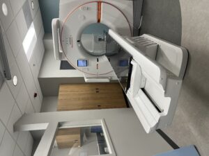 CT scanner in a medical room with patient table extended for imaging. Large circular gantry labeled Siemens Healthineers dominates right side; wooden cabinets along the wall. ceiling lights above.