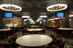 A large dining hall with round marble tables and burgundy chairs under oversized crystal chandeliers; screens line the walls.