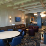 Spacious hotel lounge with round white tables, blue chairs, and purple ottomans along paneled beige walls.