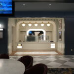 Interior of a cafe named Silks & Steam with a white counter, striped walls, and arched doorway; round marble tables with red chairs in foreground; blue walls frame the scene.
