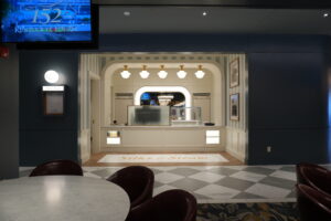 Interior of a cafe named Silks & Steam with a white counter, striped walls, and arched doorway; round marble tables with red chairs in foreground; blue walls frame the scene.