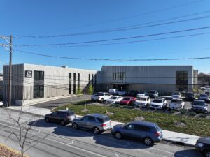 Parking lot full of cars in front of gray industrial buildings under a clear blue sky with overhead power lines across the scene.