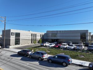 Modern white commercial building with a parking lot full of cars and overhead power lines against a clear blue sky.