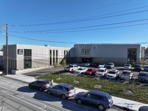 Modern industrial office building with a corrugated white facade, adjacent parking lot filled with cars under a clear blue sky and overhead power lines.