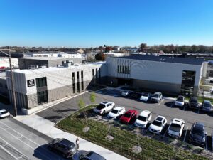 Aerial view of a modern office campus with white metal buildings, a parking lot full of cars, and a clear blue sky above.