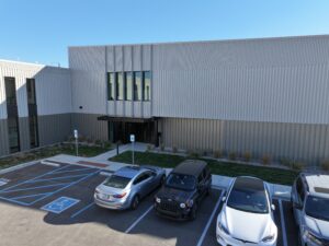 Exterior view of a modern metal-clad building with vertical panels and large windows; foreground shows a parking lot with blue handicap spaces and several cars.