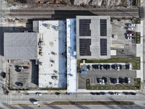 Aerial view of a commercial building complex with flat roofs, solar panels on the right roof, a parking lot with cars, and railroad tracks along the top edge.