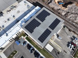 Aerial view of a flat-roofed building covered with solar panels, adjacent parking lot with cars, and railway tracks running along a rocky construction site nearby.