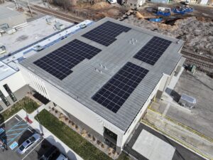 Aerial view of a commercial roof with multiple solar panels, beside railroad tracks and a construction site.