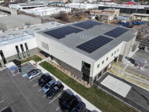 Aerial view of a grey industrial building with several large solar panel arrays on its flat roof, surrounded by a parking lot of cars and nearby rail tracks.