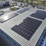 Aerial view of a flat commercial roof covered with numerous solar panels, with a parking lot and nearby street visible beyond the building edge.
