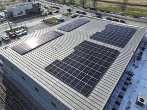 Aerial view of a flat commercial roof covered with numerous solar panels, with a parking lot and nearby street visible beyond the building edge.