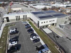 Aerial view of a modern two-story commercial building with solar panels on the roof and a parking lot with several cars.
