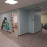 Clinic lobby with a wheelchair mural on a blue wall, curved reception desk on the right, and a seating area with a landscape painting in the background.