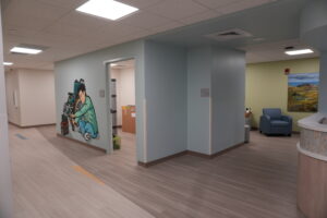 Clinic lobby with a wheelchair mural on a blue wall, curved reception desk on the right, and a seating area with a landscape painting in the background.