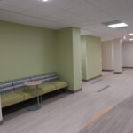 Waiting area in a clinic: lime-green walls, a long patterned upholstered bench, a small round table, and a hallway with an exit sign.