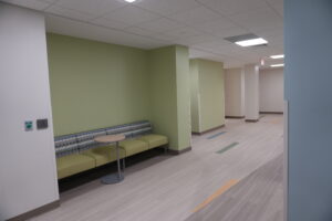 Waiting area in a clinic: lime-green walls, a long patterned upholstered bench, a small round table, and a hallway with an exit sign.