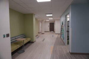 Wide hospital-like hallway with green seating alcove on left, mural on right wall, and door at far end under fluorescent ceiling lights.