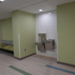 Lobby area with lime-green walls, white tiled wall housing a dual drinking fountain, and a seating bench along the left; a person stands near a doorway in the background.