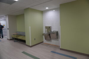 Lobby area with lime-green walls, white tiled wall housing a dual drinking fountain, and a seating bench along the left; a person stands near a doorway in the background.