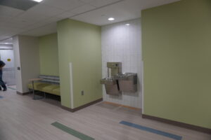 Interior hallway with pale green walls, a white tiled column and dual drinking fountain; bench seating along the left wall.