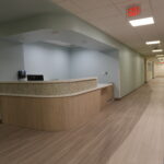 Reception desk in a modern hallway with light blue walls and wood-toned counters, illuminated by ceiling lights and an exit sign overhead.