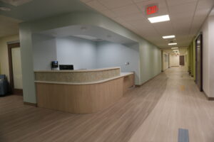 Reception desk in a modern hallway with light blue walls and wood-toned counters, illuminated by ceiling lights and an exit sign overhead.